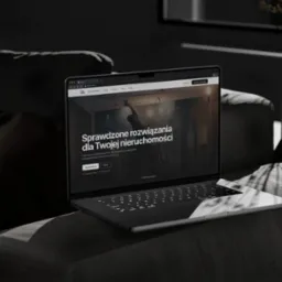 Laptop na ciemnoszarym fotelu prezentuje stronę internetową firmy budowlanej z hasłem 'Sprawdzone rozwiązania dla Twojej nieruchomości', w tle wazon z kwiatami.