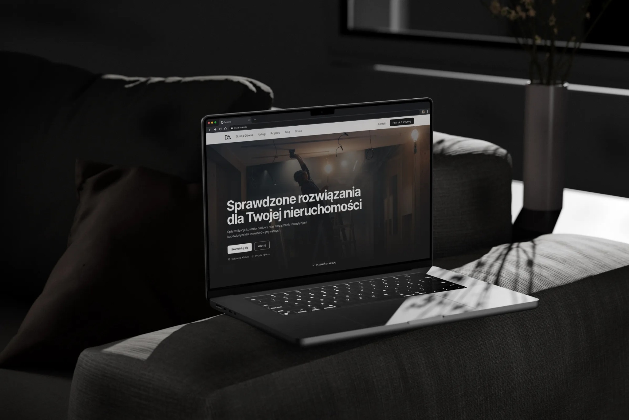 Laptop na ciemnoszarym fotelu prezentuje stronę internetową firmy budowlanej z hasłem 'Sprawdzone rozwiązania dla Twojej nieruchomości', w tle wazon z kwiatami.