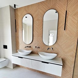 STOLRYS Meble łazienkowe na wymiar - Nowoczesna łazienka z dwiema owalnymi umywalkami nablatowymi, białym blatem z frezowanymi frontami, dwoma owalnymi lustrami w czarnych ramach i drewnianą ścianą z geometrycznym wzorem.