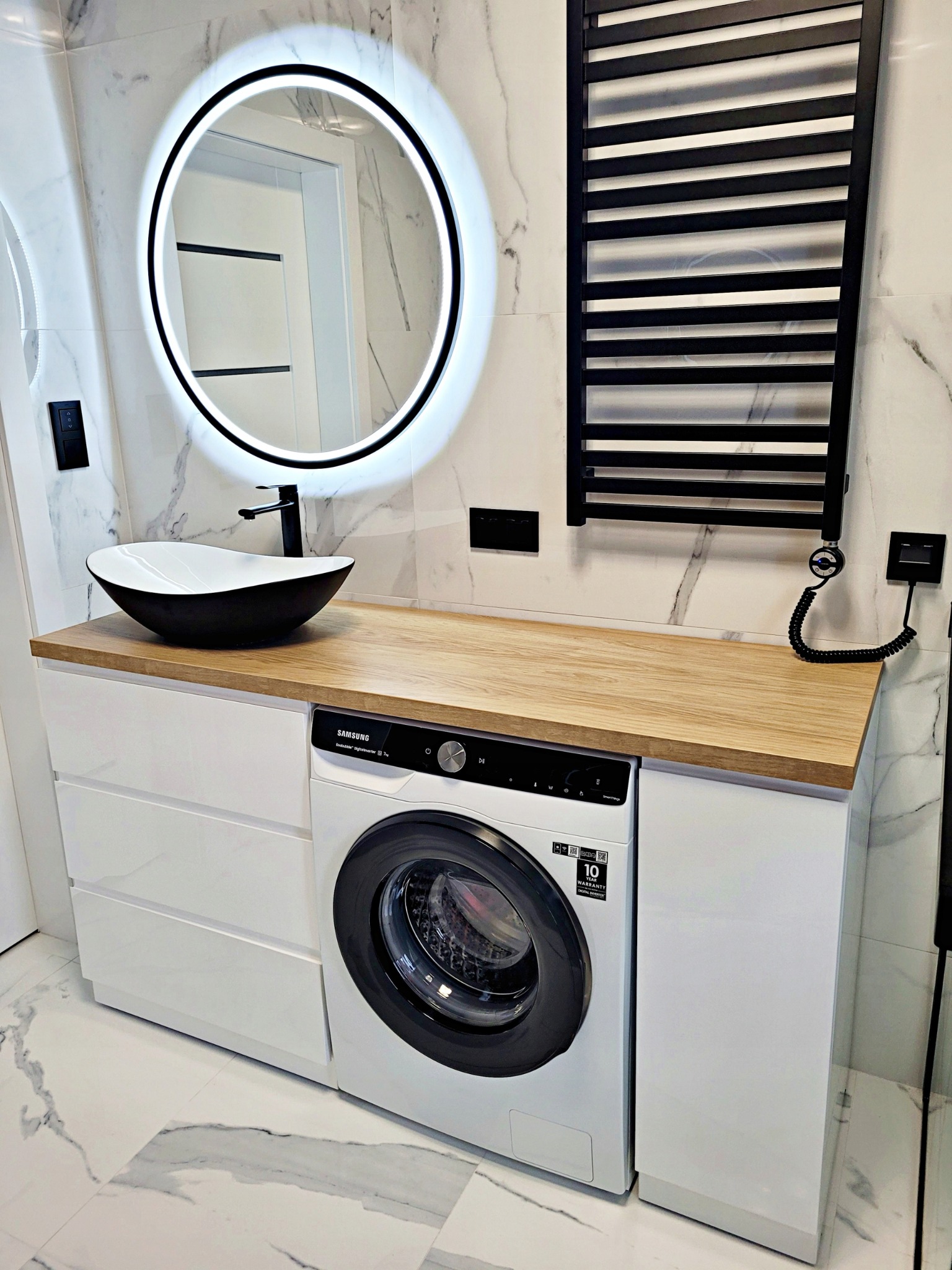 Nowoczesna łazienka z białą szafką pod umywalkę z drewnianym blatem, na którym stoi czarno-biała umywalka i bateria, obok pralka zabudowana w szafce, okrągłe lustro z podświetleniem LED i czarny...