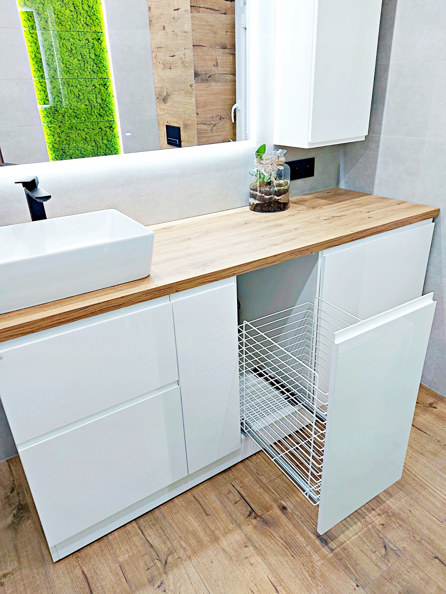 Biała szafka łazienkowa z drewnianym blatem, wysuwanym metalowym koszem na pranie i prostokątną umywalką nablatową, uzupełniona dekoracyjną kompozycją roślinną w szklanym słoju.