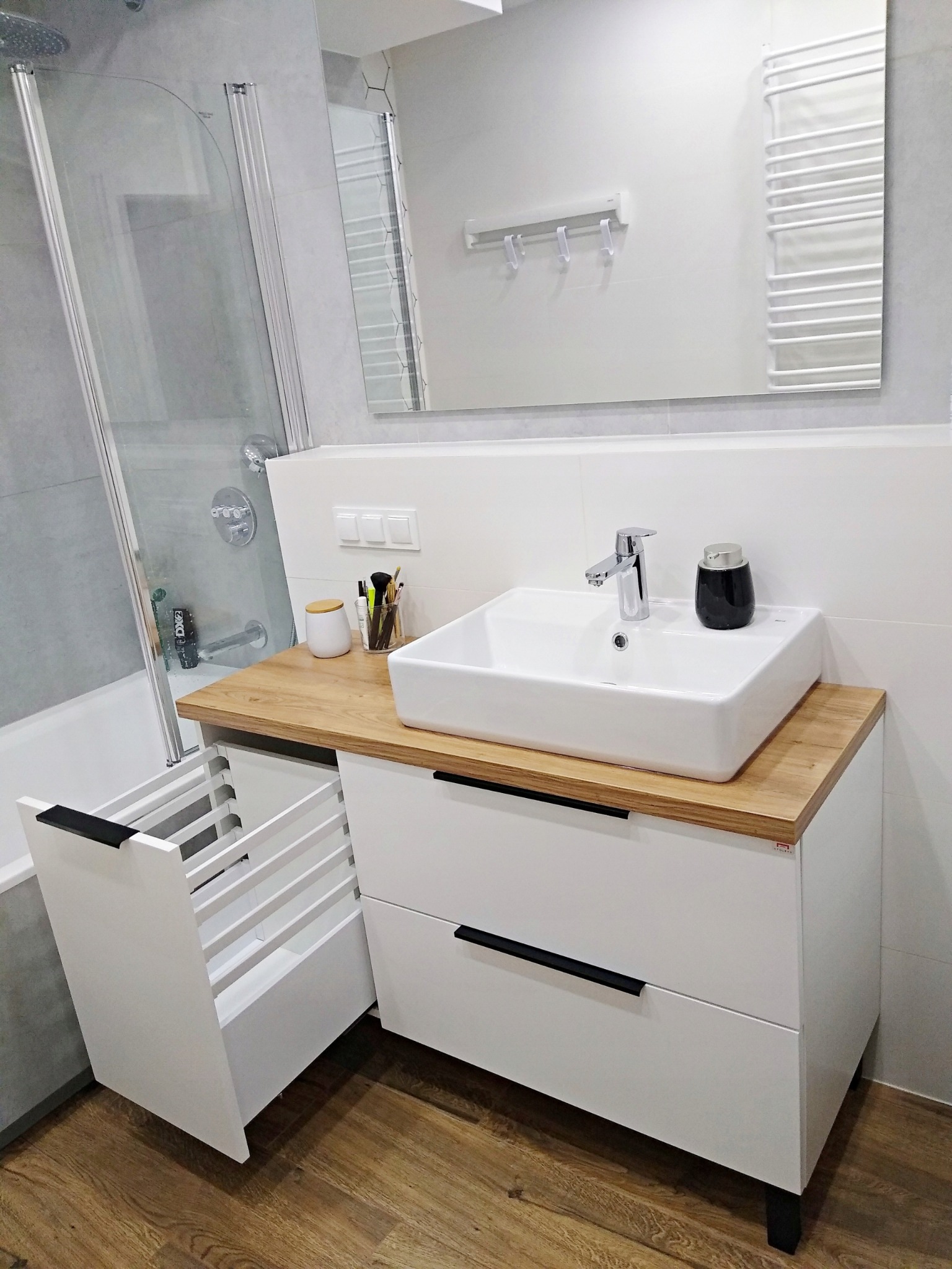 Nowoczesna łazienka z białą szafką pod umywalkę z drewnianym blatem i wysuwanym koszem na pranie, widoczna bateria umywalkowa i dozownik mydła, w tle kabina prysznicowa i grzejnik drabinkowy.