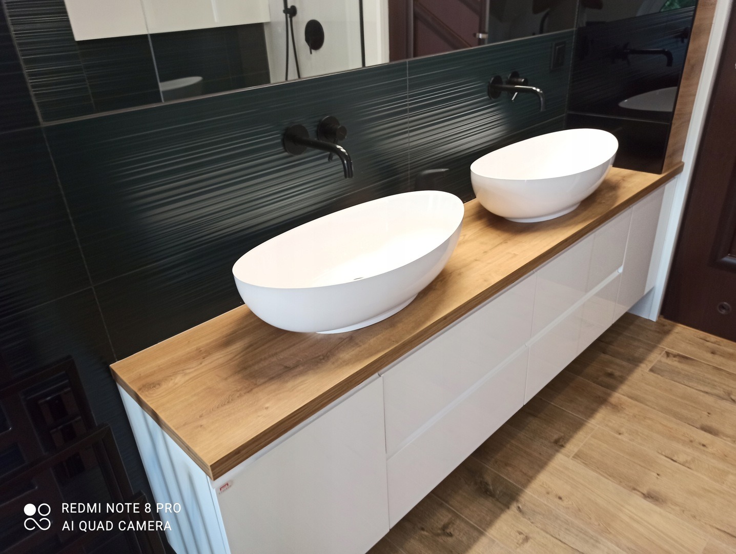 Łazienka z dwoma owalnymi umywalkami nablatowymi, blatem drewnianym i szafką z białymi frontami, na tle ciemnozielonych płytek i czarnej armatury.