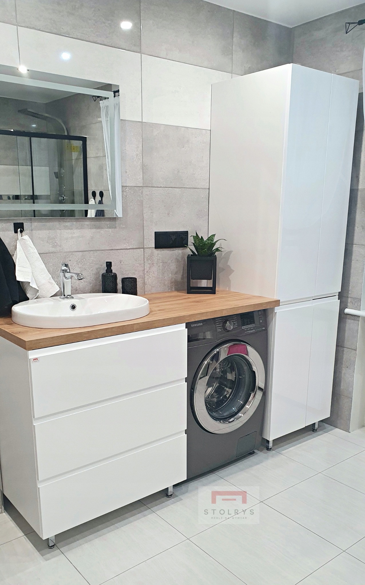 Nowoczesna łazienka z drewnianym blatem, białą umywalką nablatową, szafką z szufladami, pralką Samsung i wysoką białą szafką, całość w szaro-białej tonacji z elementami dekoracyjnymi.