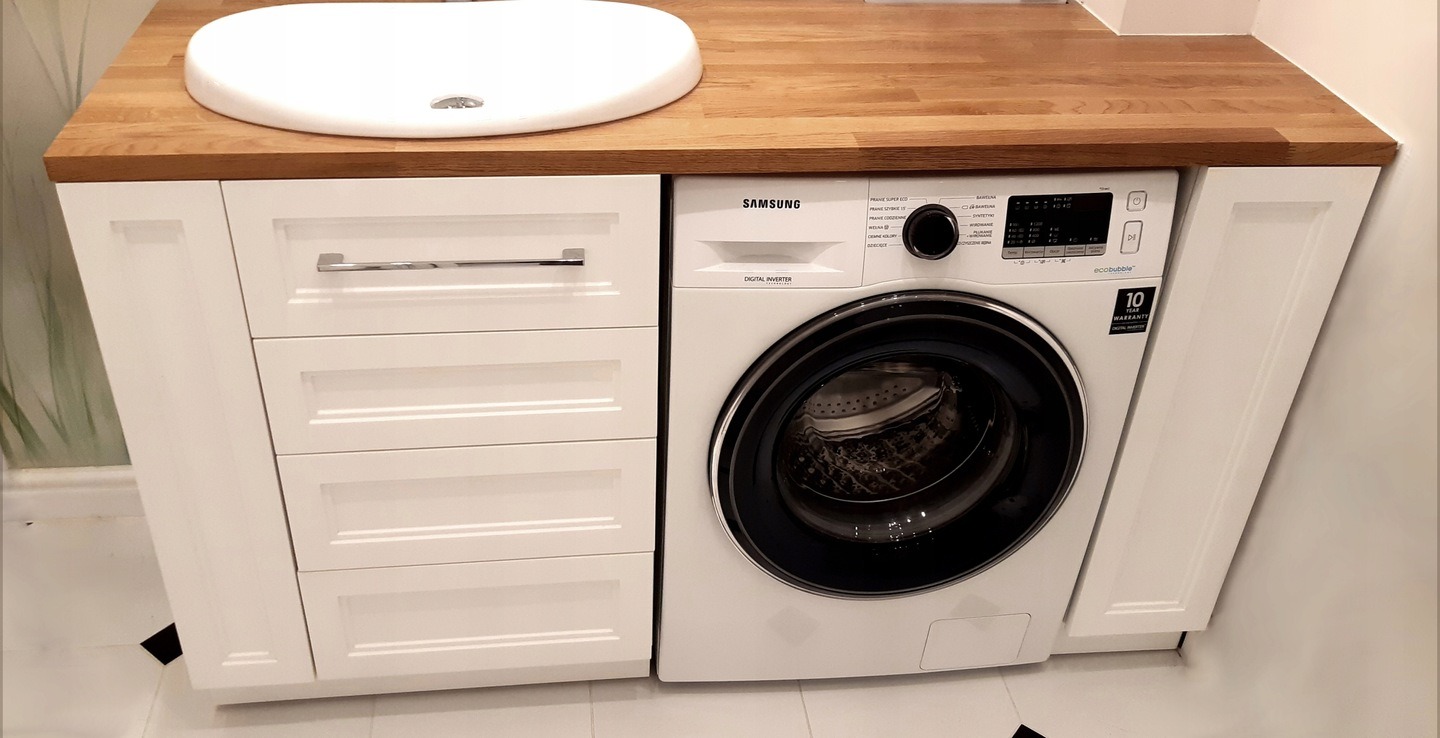 Łazienka z umywalką nablatową na drewnianym blacie, białe szafki z szufladami i pralka w zabudowie.