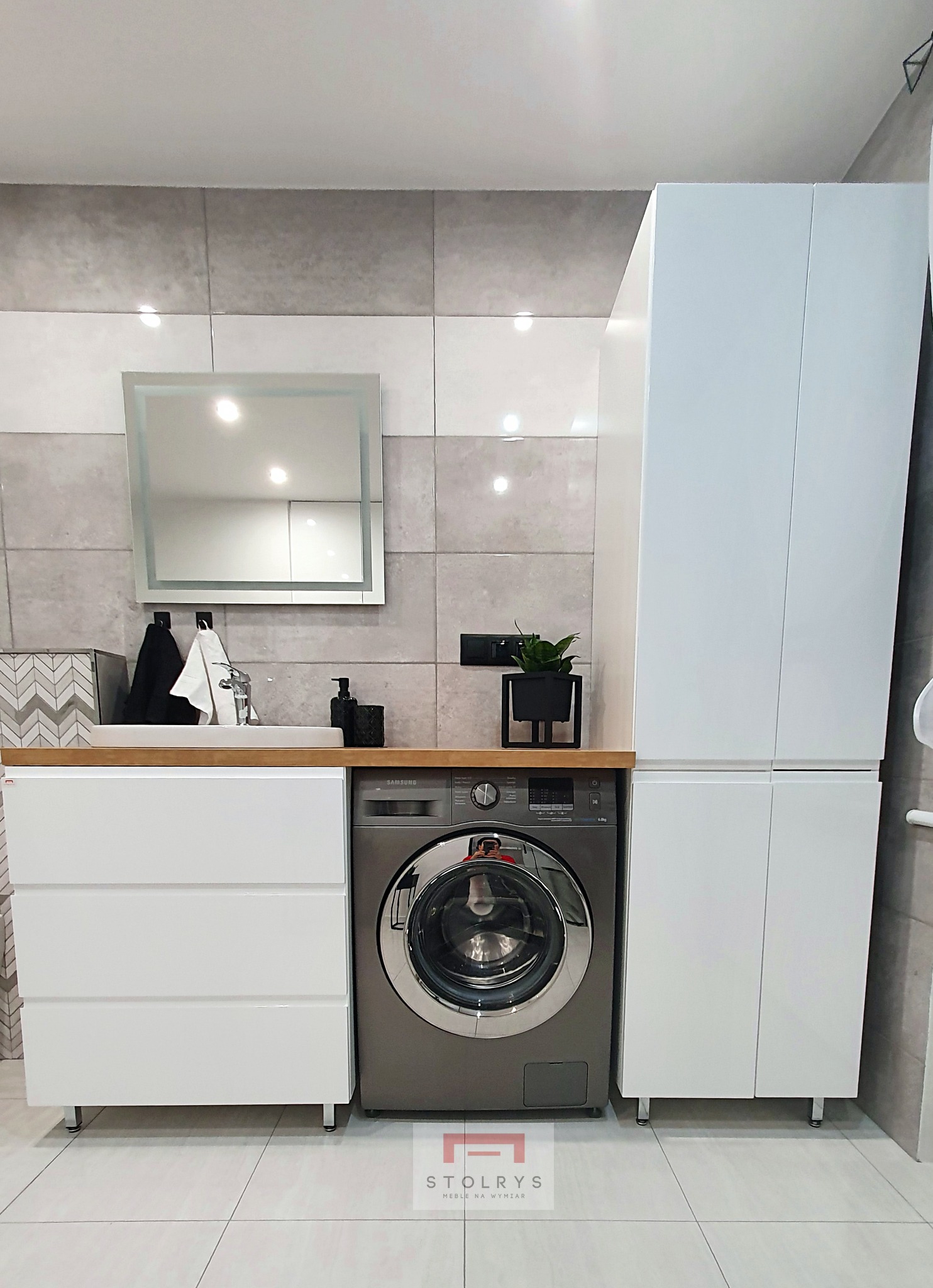 Nowoczesna łazienka z zabudową meblową: białe szafki z szufladami pod drewnianym blatem, pralka w kolorze grafitowym, wysoka szafka z białymi frontami, lustro z podświetleniem i szare płytki...