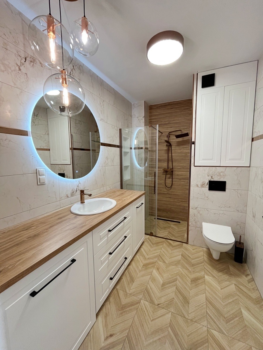 Nowoczesna łazienka z drewnianym blatem pod umywalką, oświetlonym lustrem i kabiną prysznicową z drewnianymi panelami, wykończona płytkami imitującymi marmur i drewno, oświetlona wiszącymi lampami...