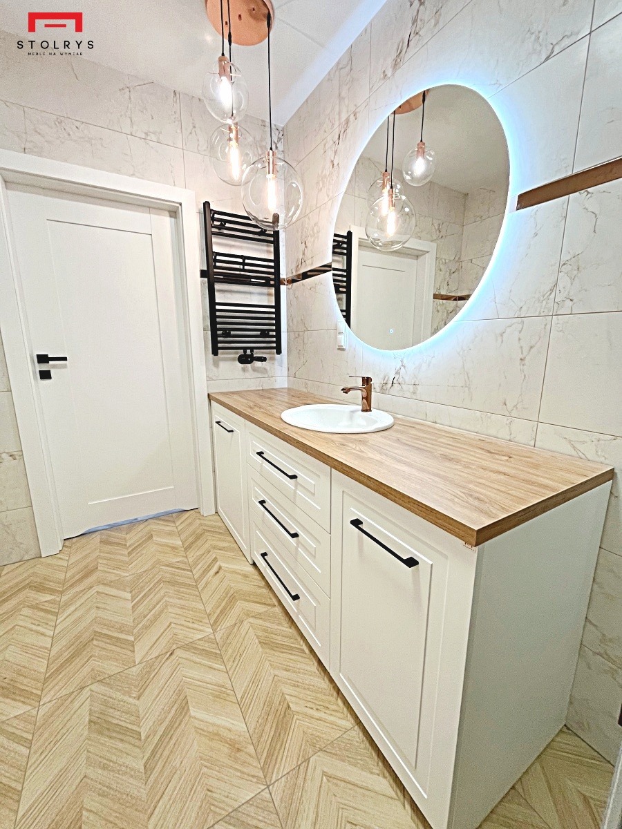 Biała szafka łazienkowa z drewnianym blatem, umywalką i podświetlanym lustrem w owalnej ramie, widoczna w łazience w Nowym Mieście Lubawskim z marmurowymi płytkami i wzorzystą podłogą ułożoną...