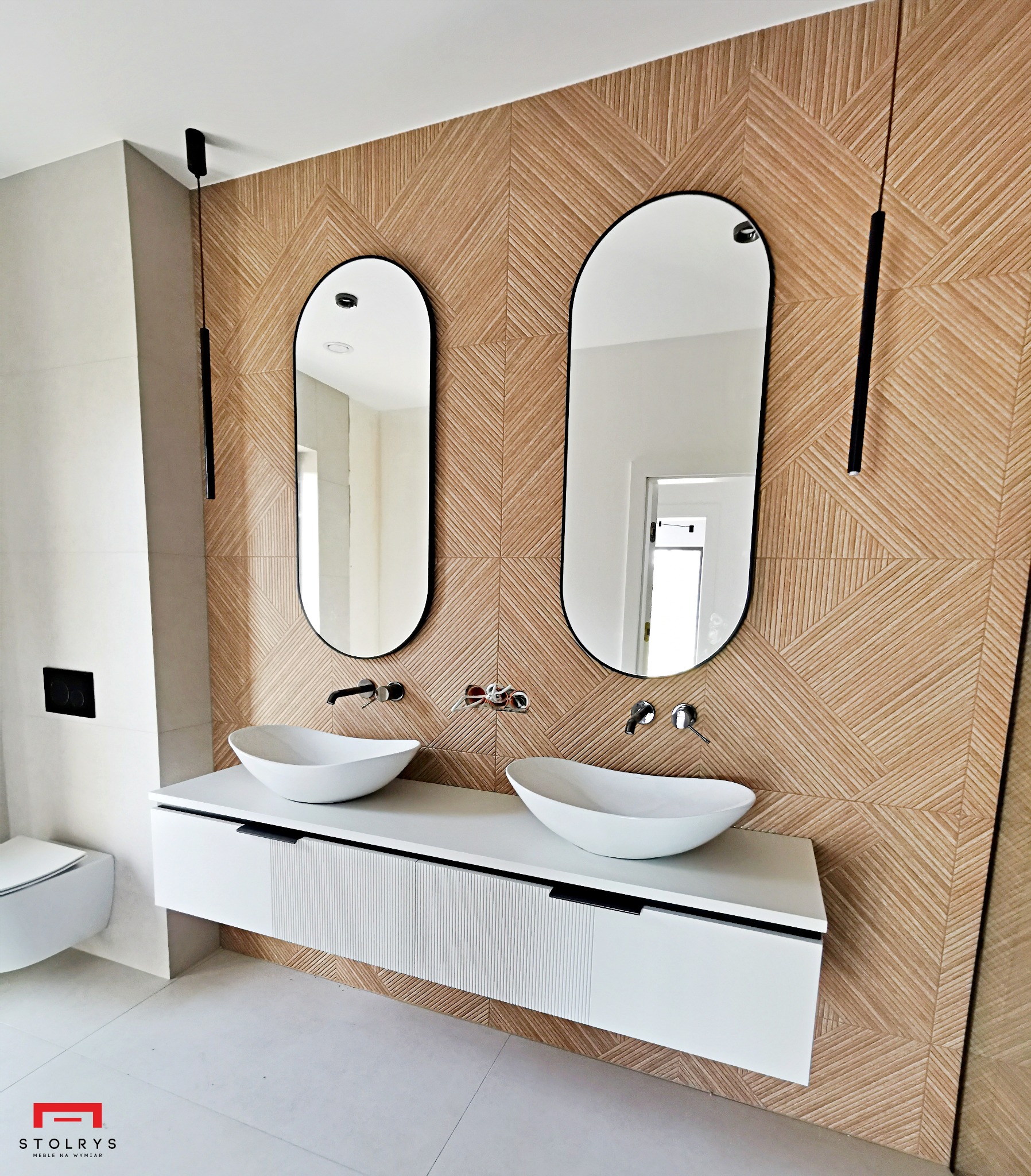 Nowoczesna łazienka z dwiema owalnymi umywalkami nablatowymi, białym blatem z frezowanymi frontami, dwoma owalnymi lustrami w czarnych ramach i drewnianą ścianą z geometrycznym wzorem.