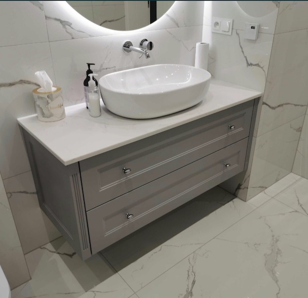 Szara szafka łazienkowa z dwiema szufladami, białym blatem i owalną umywalką nablatową, na tle marmurowych płytek i okrągłego lustra z podświetleniem LED.