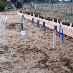 TOBIASZ CYBULSKI - Przygotowanie terenu pod instalacje: widoczne rury kanalizacyjne wystające z ziemi, obok drewniane szalunki na fundamencie, w tle dom jednorodzinny i ogrodzenie z siatki.