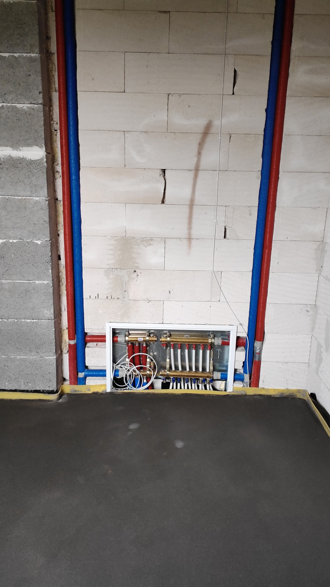 Rozdzielacz ogrzewania podłogowego w białej obudowie, widoczne rury w izolacji czerwonej i niebieskiej, ściany z betonu komórkowego i wylewka podłogowa w surowym stanie.