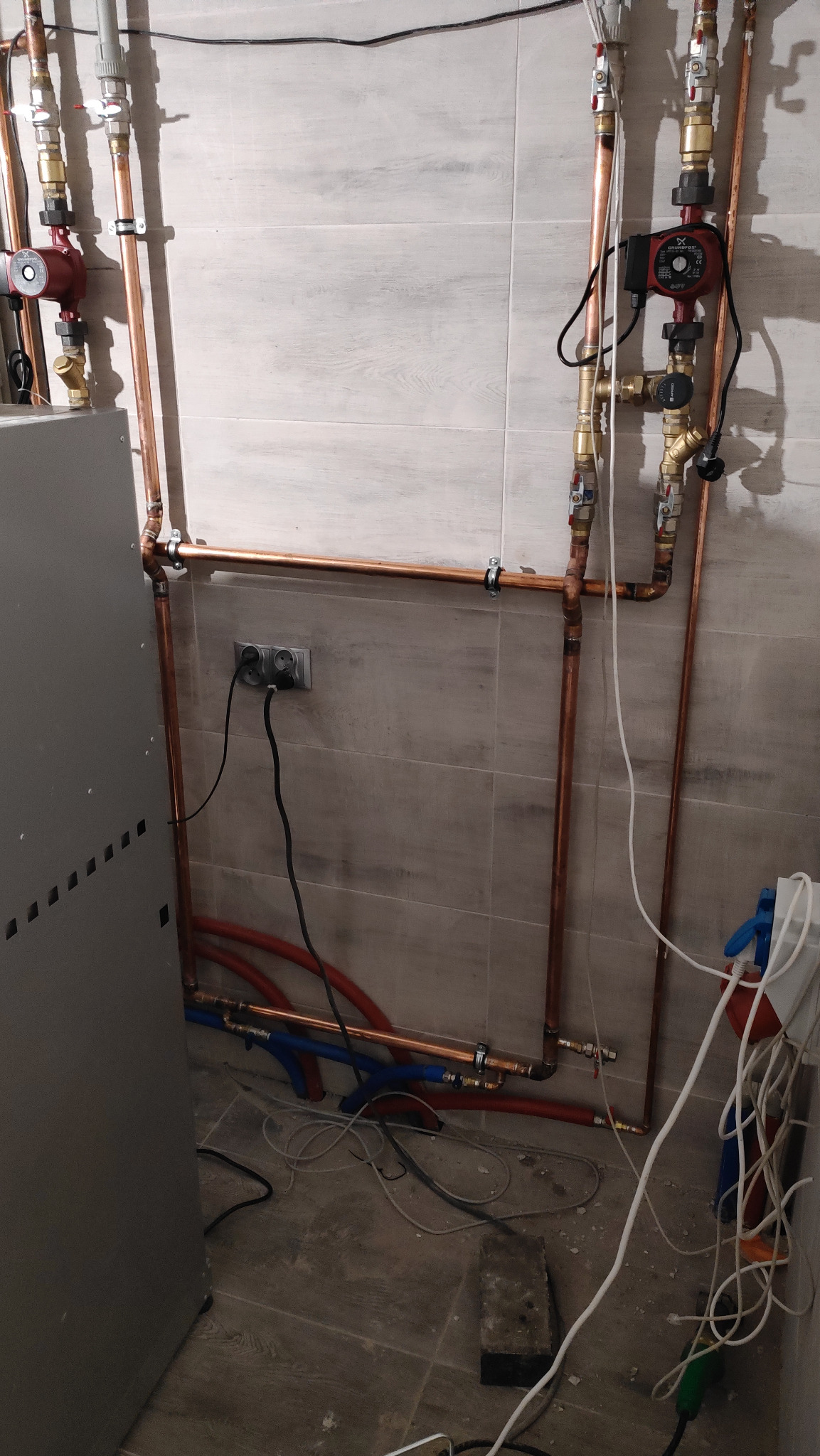 Miedziane rury instalacji grzewczej z pompami obiegowymi, zaworami i podłączeniem elektrycznym na tle szarej płytki ściennej, widoczne także fragmenty niebieskich i czerwonych rur oraz kabli...