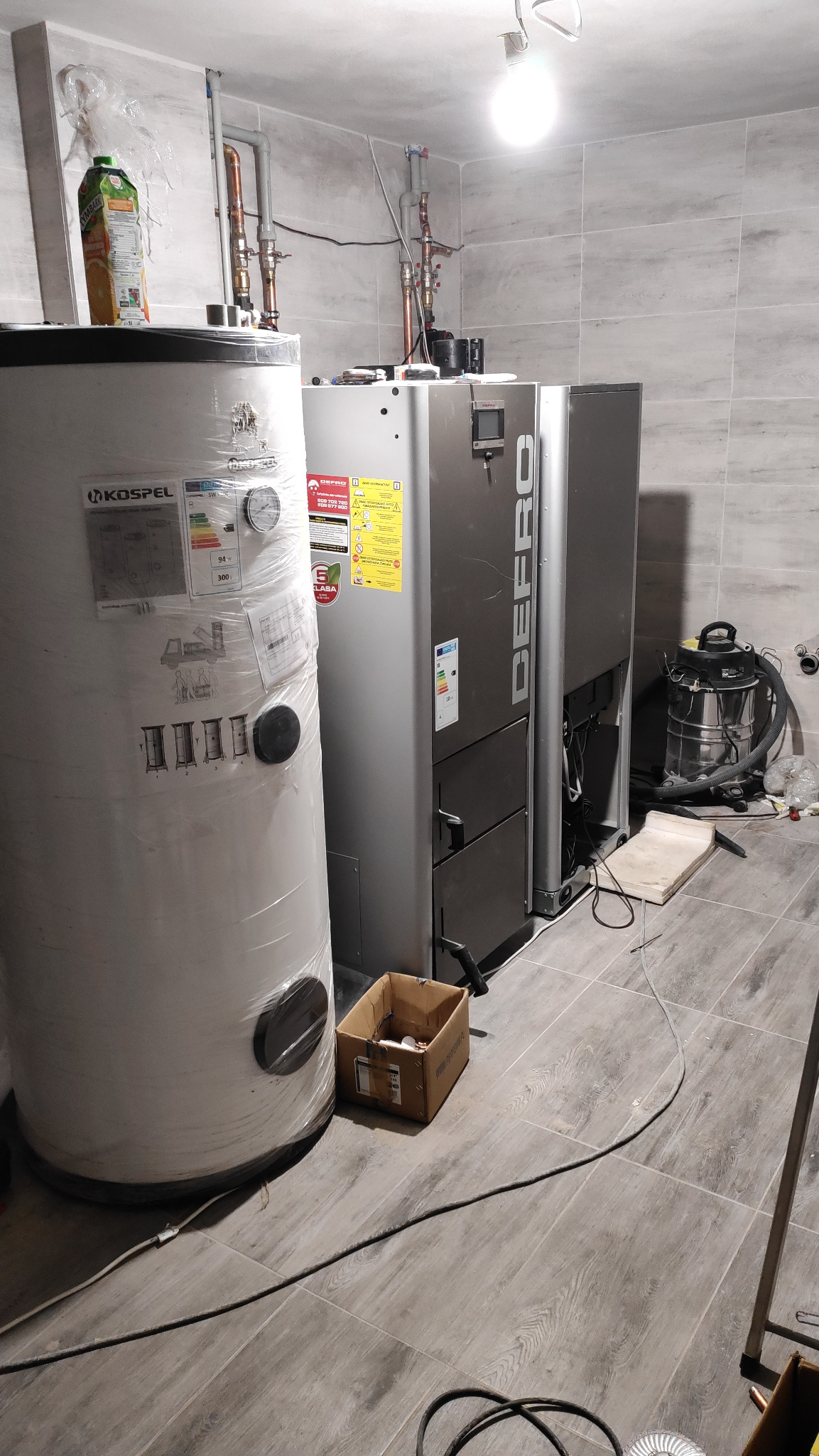 Nowoczesny piec CO marki Defro obok owiniętego folią ochronną zasobnika ciepłej wody Kospel w pomieszczeniu z szarą płytką imitującą drewno, widoczny odkurzacz przemysłowy i kartonowe pudełko.