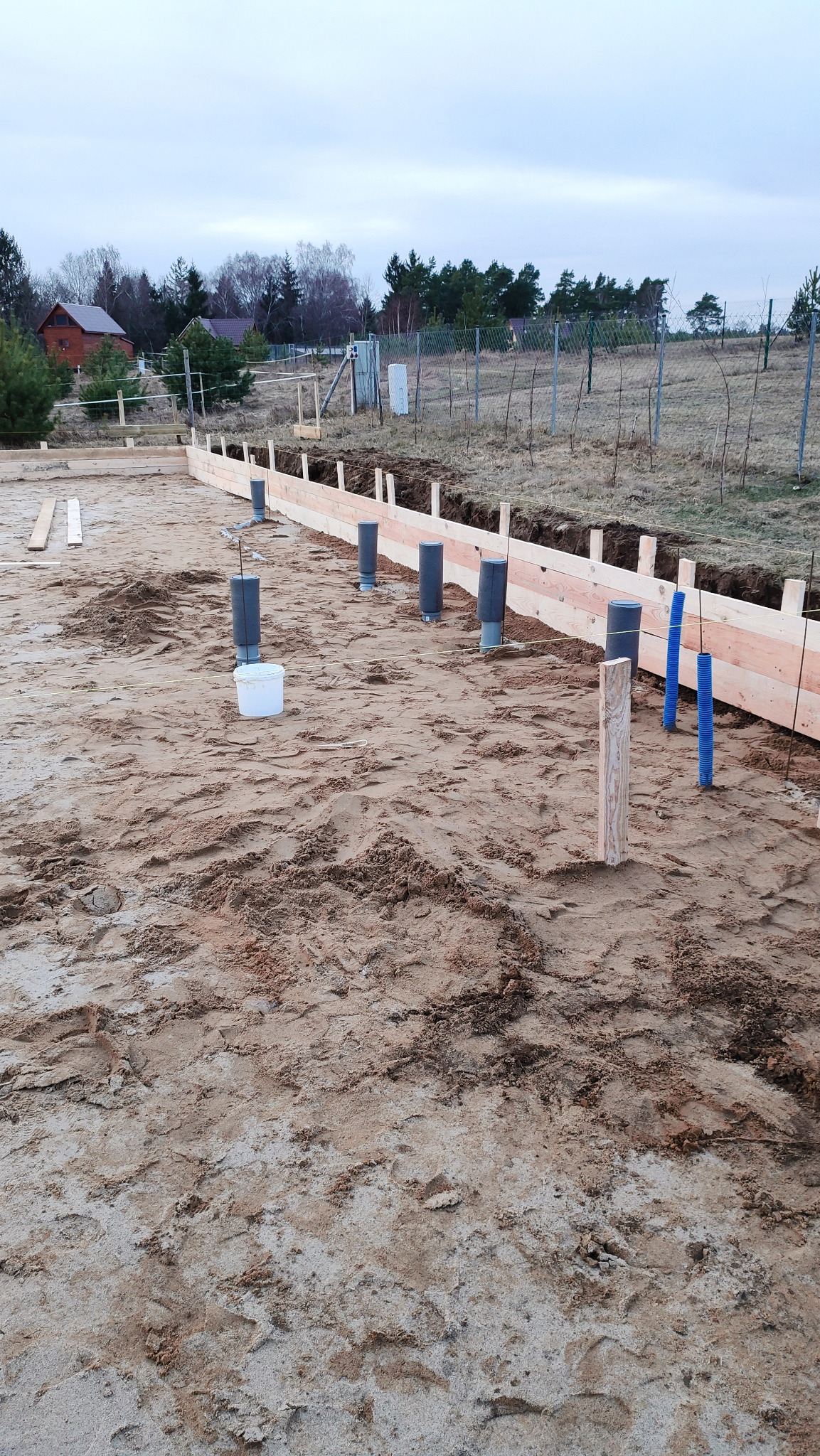 Przygotowanie terenu pod instalacje: widoczne rury kanalizacyjne wystające z ziemi, obok drewniane szalunki na fundamencie, w tle dom jednorodzinny i ogrodzenie z siatki.