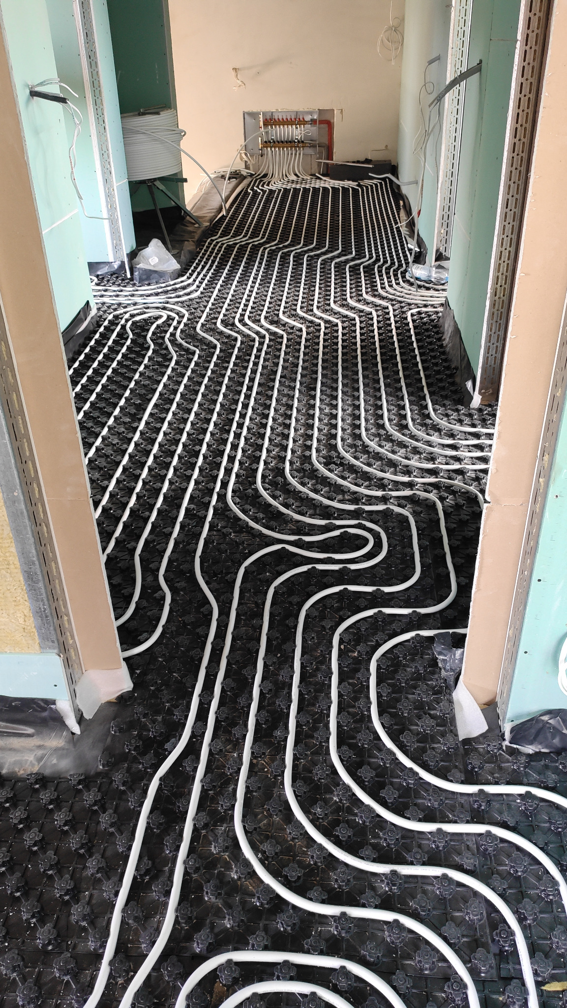 Rozłożone białe rury ogrzewania podłogowego na czarnej macie z mocowaniami, widok z góry na instalację w trakcie prac wykończeniowych.