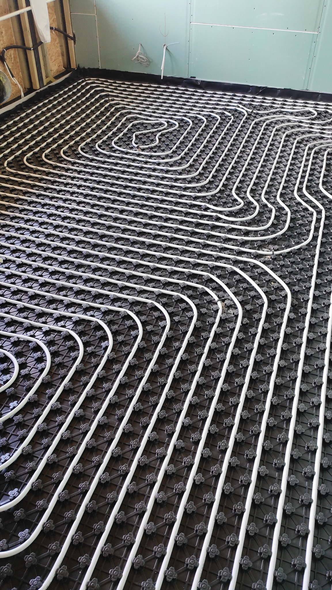 Białe rury ogrzewania podłogowego ułożone w pętle na czarnej, modułowej macie montażowej, widok z góry na surową podłogę w trakcie instalacji.