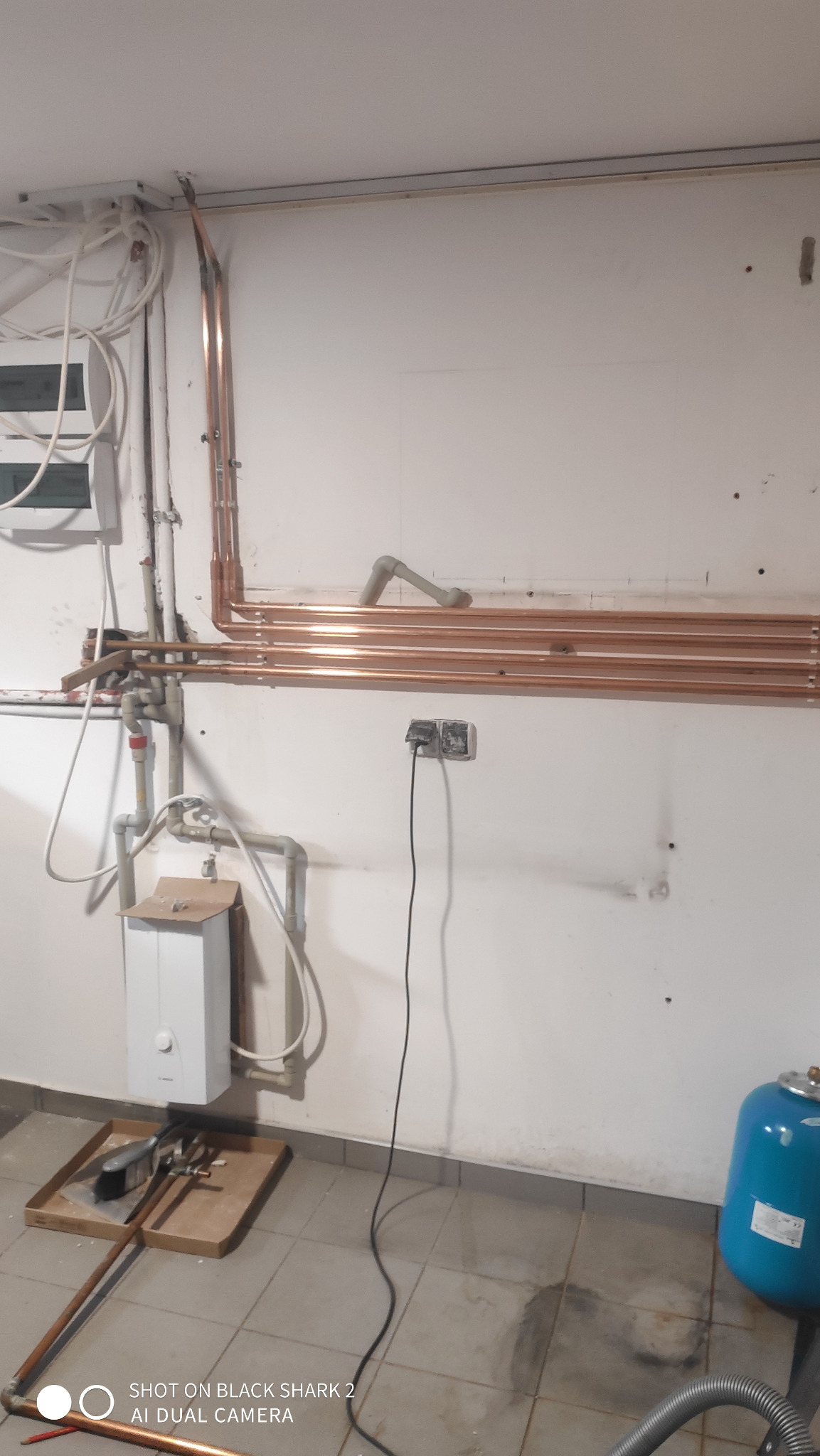 Montaż instalacji centralnego ogrzewania: miedziane rury rozprowadzone wzdłuż ściany, podłączony piec, niebieski zbiornik wyrównawczy, narzędzia na tacy na podłodze, widoczne ślady po wierceniu...