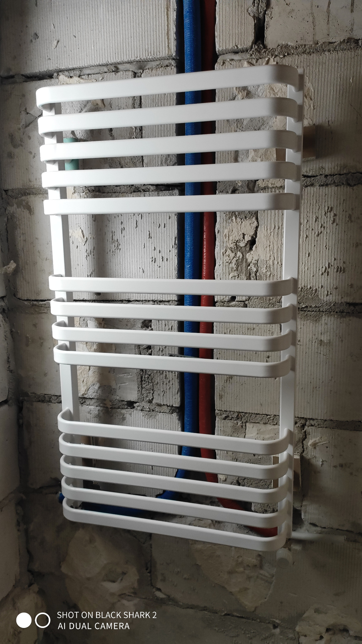 Biały grzejnik łazienkowy drabinkowy zamontowany na ścianie z surowej cegły, z widocznymi niebieskimi i czerwonymi rurami doprowadzającymi wodę.