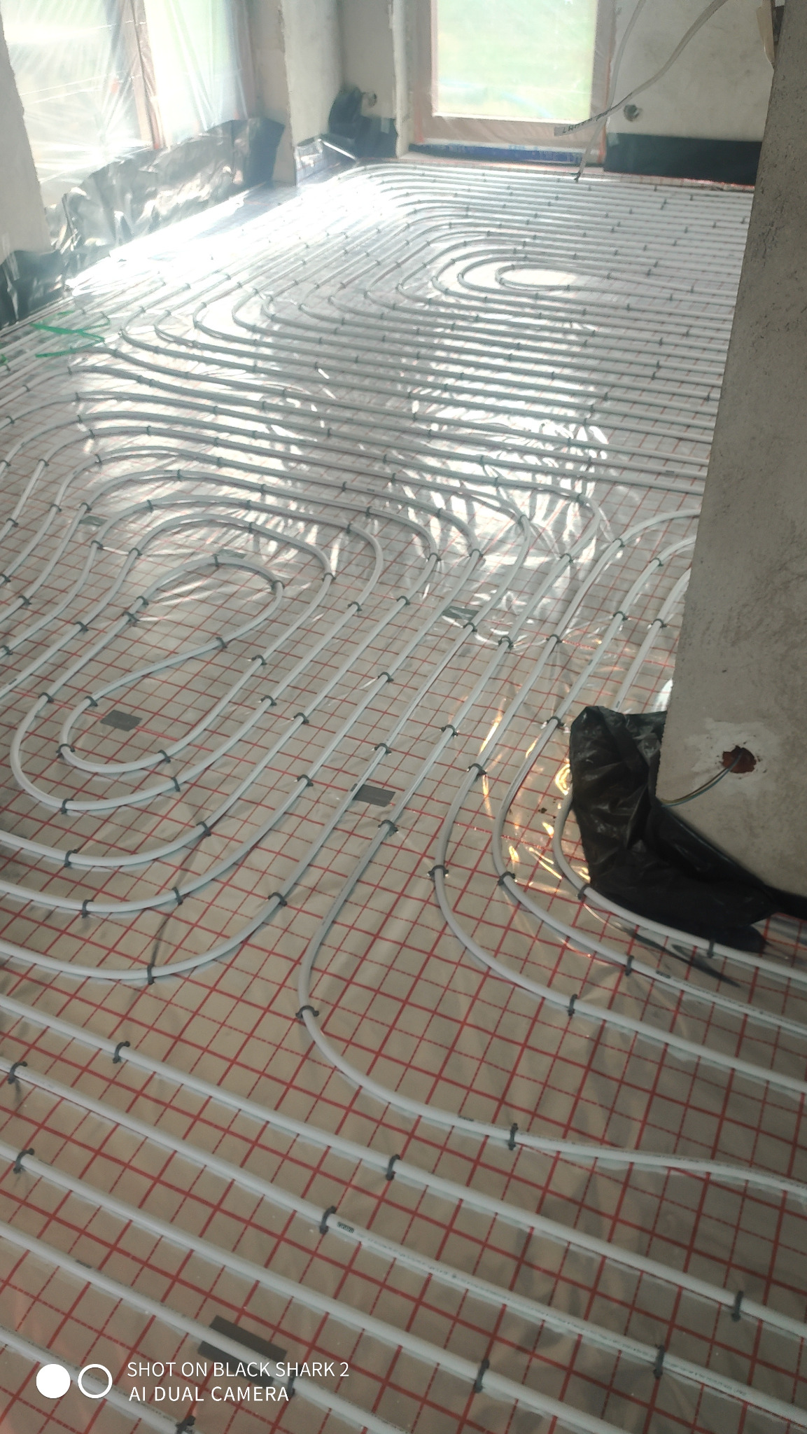 Układanie białych rur ogrzewania podłogowego na folii z czerwoną siatką, widoczne mocowania i fragment ściany z instalacją elektryczną.