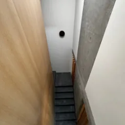 Nowoczesne schody z czarnymi stopniami, drewnianą ścianą z widocznym usłojeniem, betonową ścianą z charakterystycznymi otworami po szalunkach i okrągłym otworem wentylacyjnym w suficie.