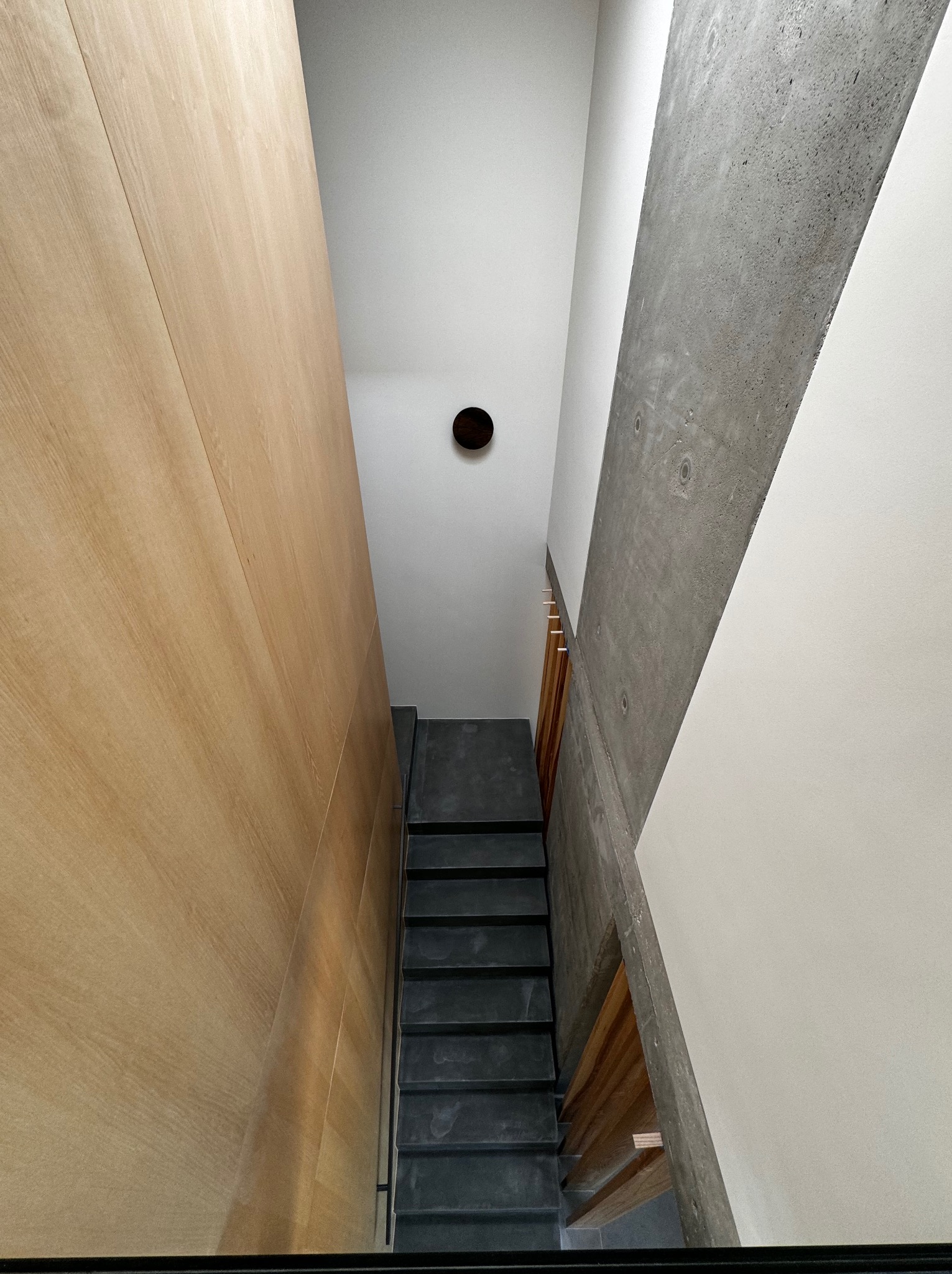 Wysokie wnętrze z betonową ścianą, schodami z ciemnego kamienia i drewnianą okładziną ścienną w ciepłym odcieniu, widok z góry na dół.