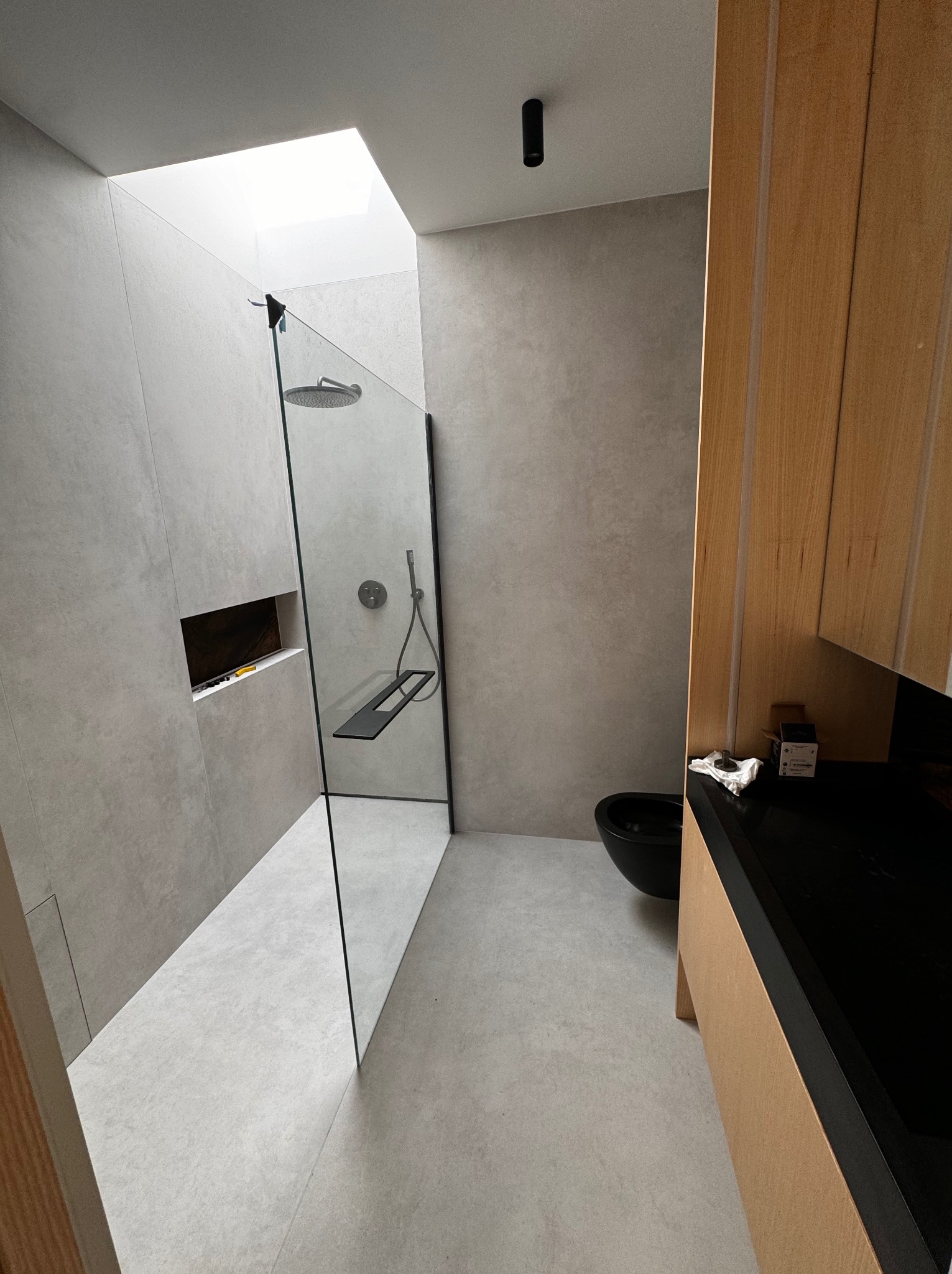 Nowoczesna łazienka z kabiną prysznicową typu walk-in, wykończona w szarościach, z drewnianą szafką pod umywalką i oknem dachowym wpadającym naturalne światło. Widoczna czarna umywalka nablatowa...