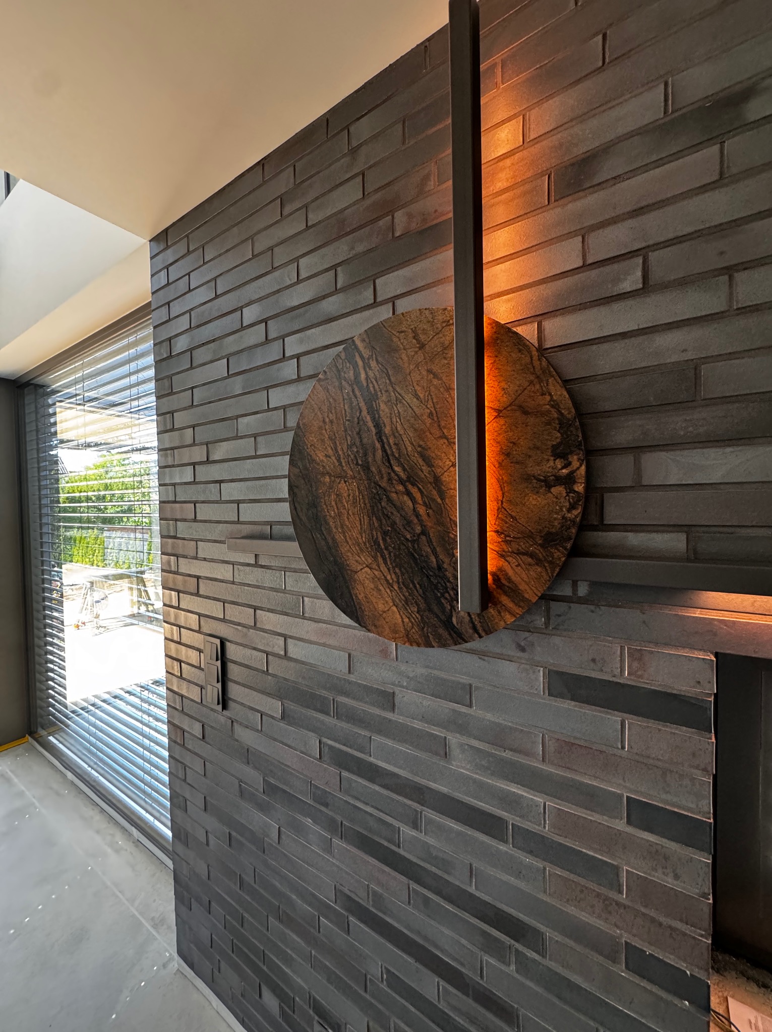 Dekoracyjna ściana z ciemnej cegły z okrągłym, marmurkowym elementem i podświetleniem LED, widoczne żaluzje okienne i fragment surowej podłogi.