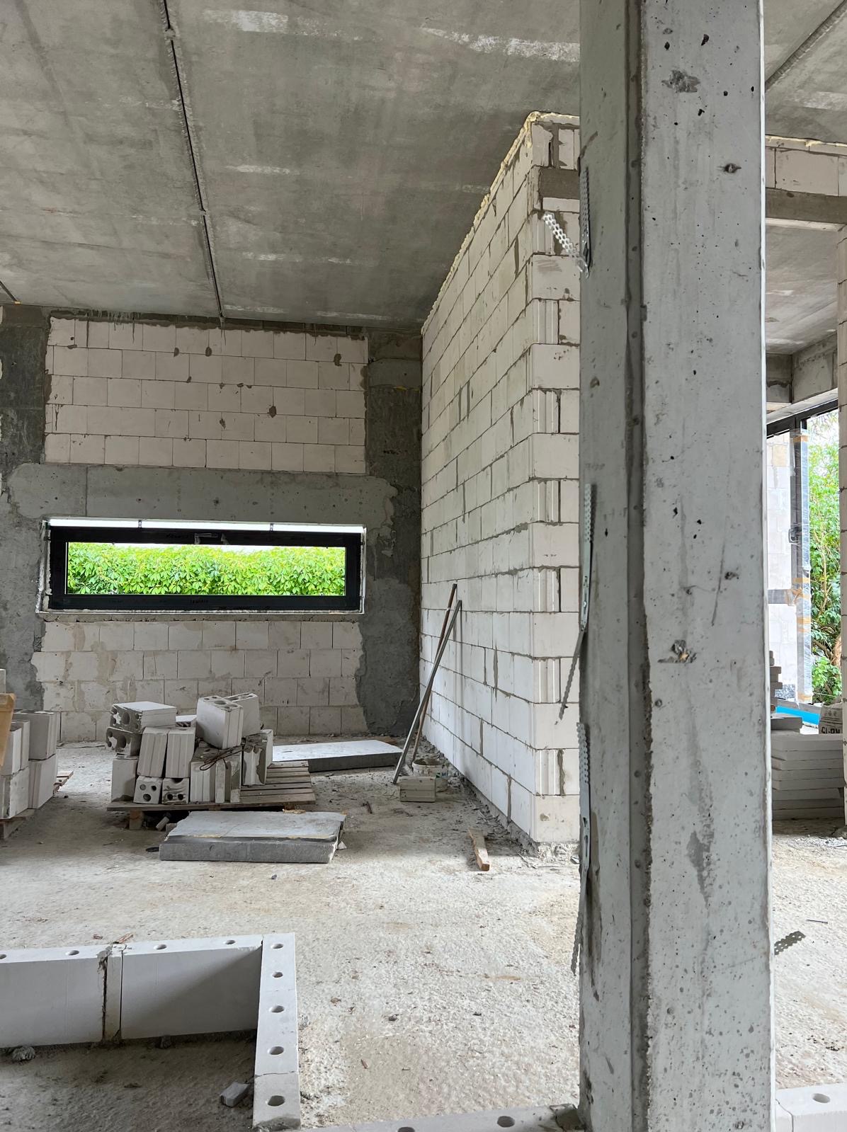 Wnętrze surowego domu w trakcie budowy: ściany z białych bloczków, okno z widokiem na zieleń, betonowy filar na pierwszym planie, paleta z bloczkami i narzędzia budowlane.