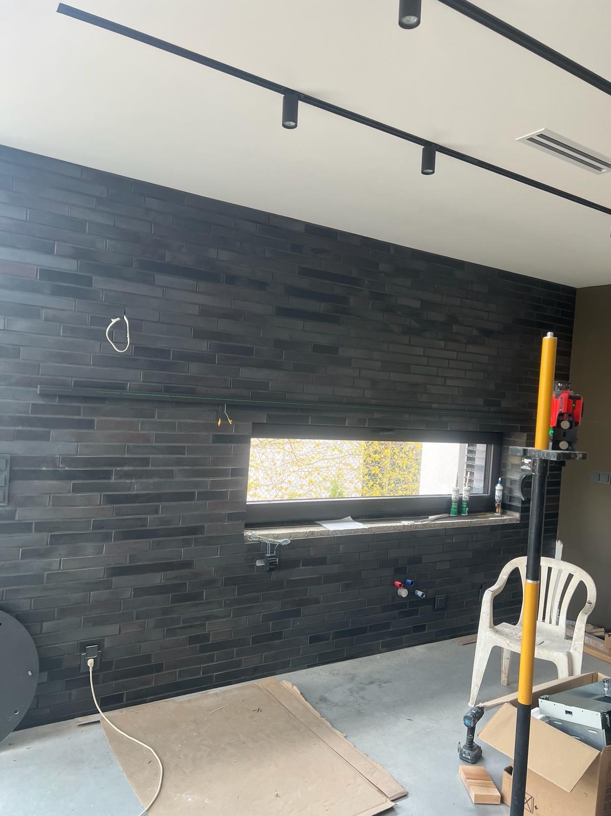 Ściana wykończona czarną, cegiełkową płytką, widoczne przewody elektryczne, okno z kamiennym parapetem, poziomica laserowa na statywie, w tle białe krzesło i kartony.