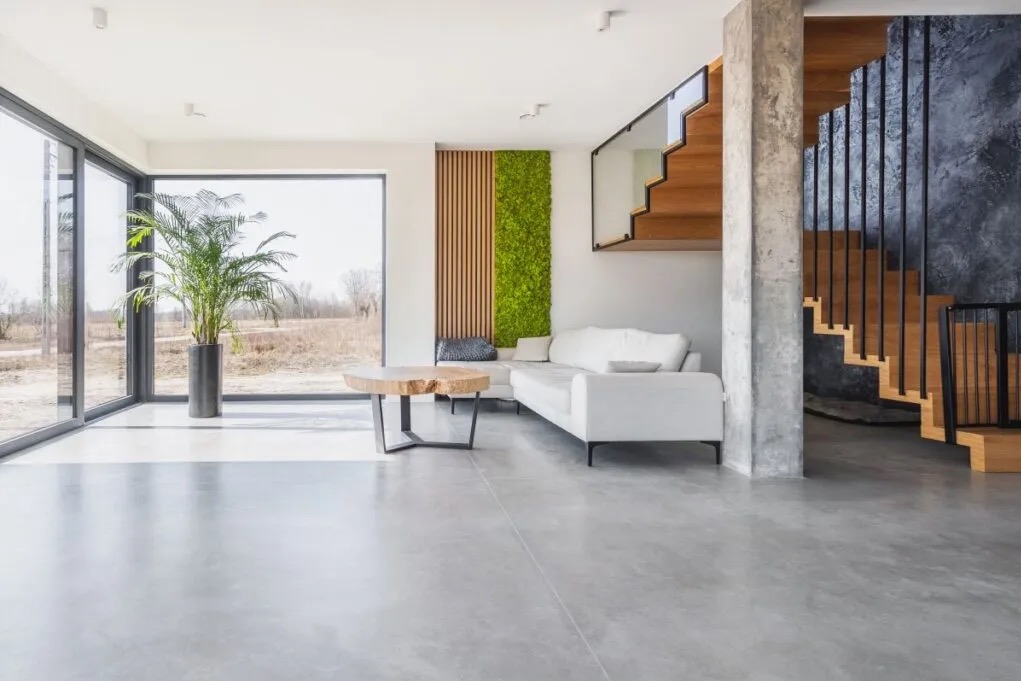 Jasne, minimalistyczne wnętrze z betonową podłogą, drewnianymi schodami z czarną balustradą, narożną sofą w kolorze złamanej bieli, stolikiem kawowym z blatem z surowego drewna i ścianą z panelami...