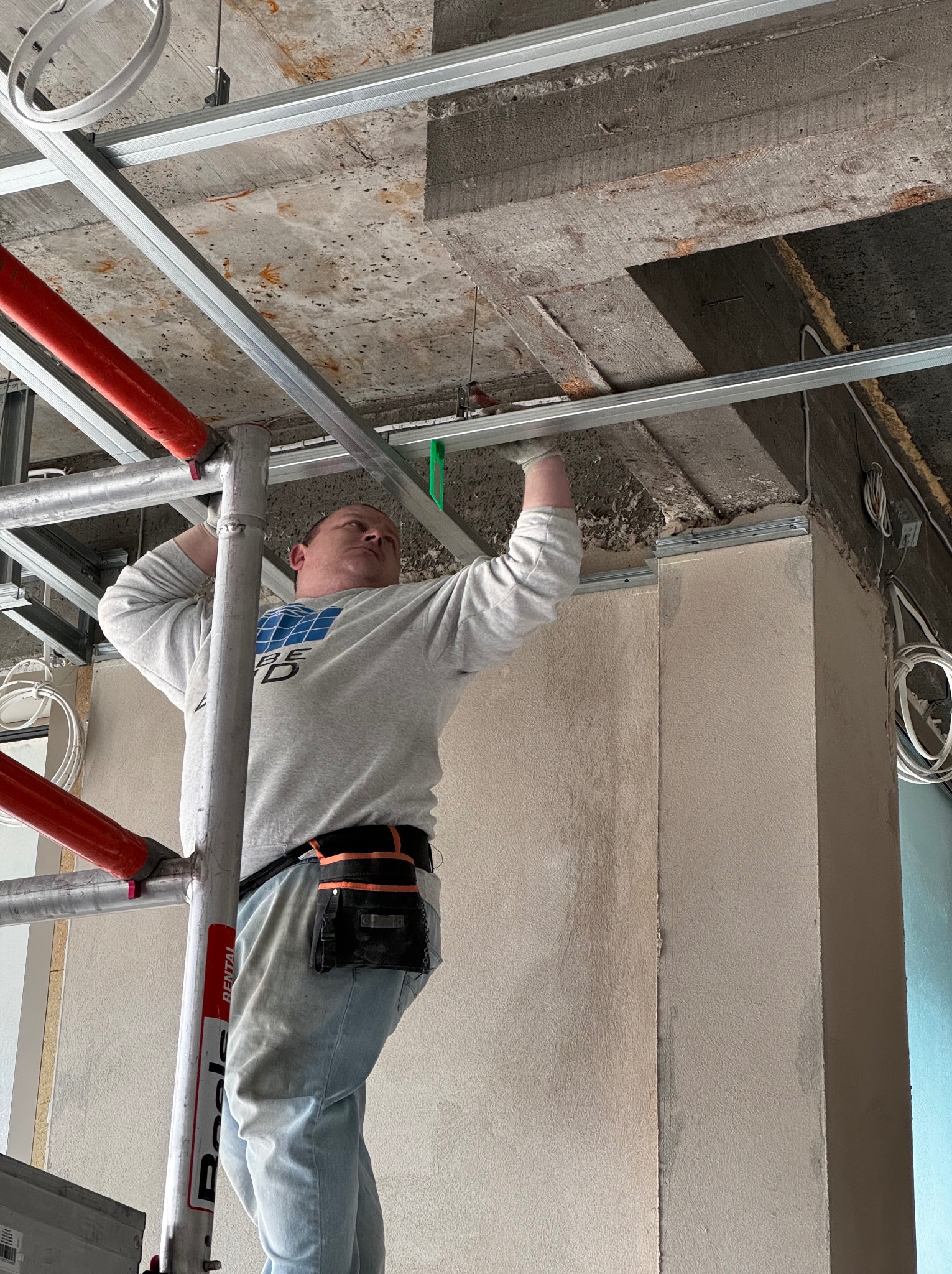 Pracownik na rusztowaniu montuje metalowy stelaż sufitu podwieszanego w pomieszczeniu w trakcie remontu, widoczne instalacje pod stropem.