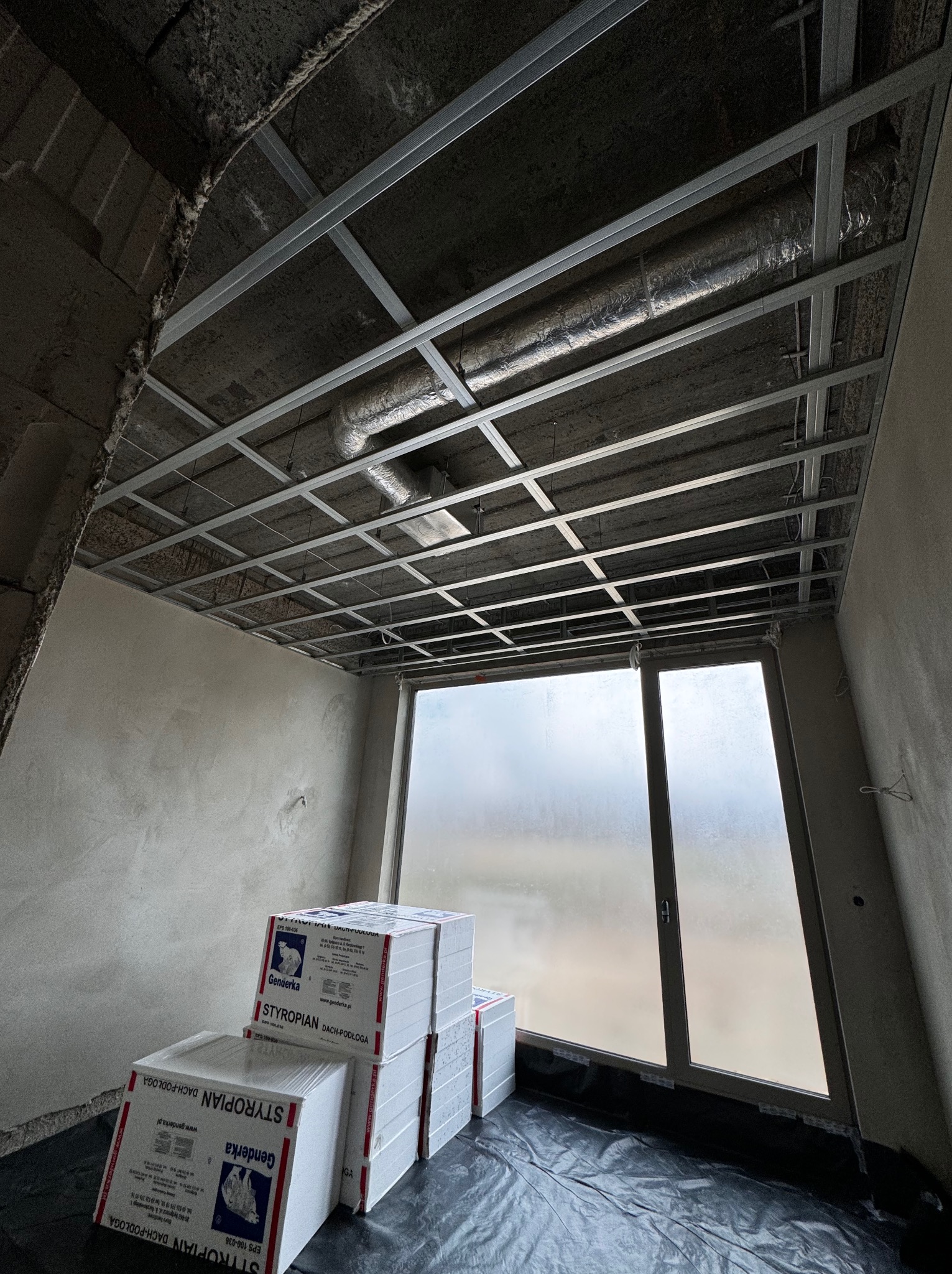 Wnętrze w trakcie remontu: metalowa konstrukcja sufitu podwieszanego, rury wentylacyjne, okno z matową szybą i stos paczek styropianu na czarnej folii.