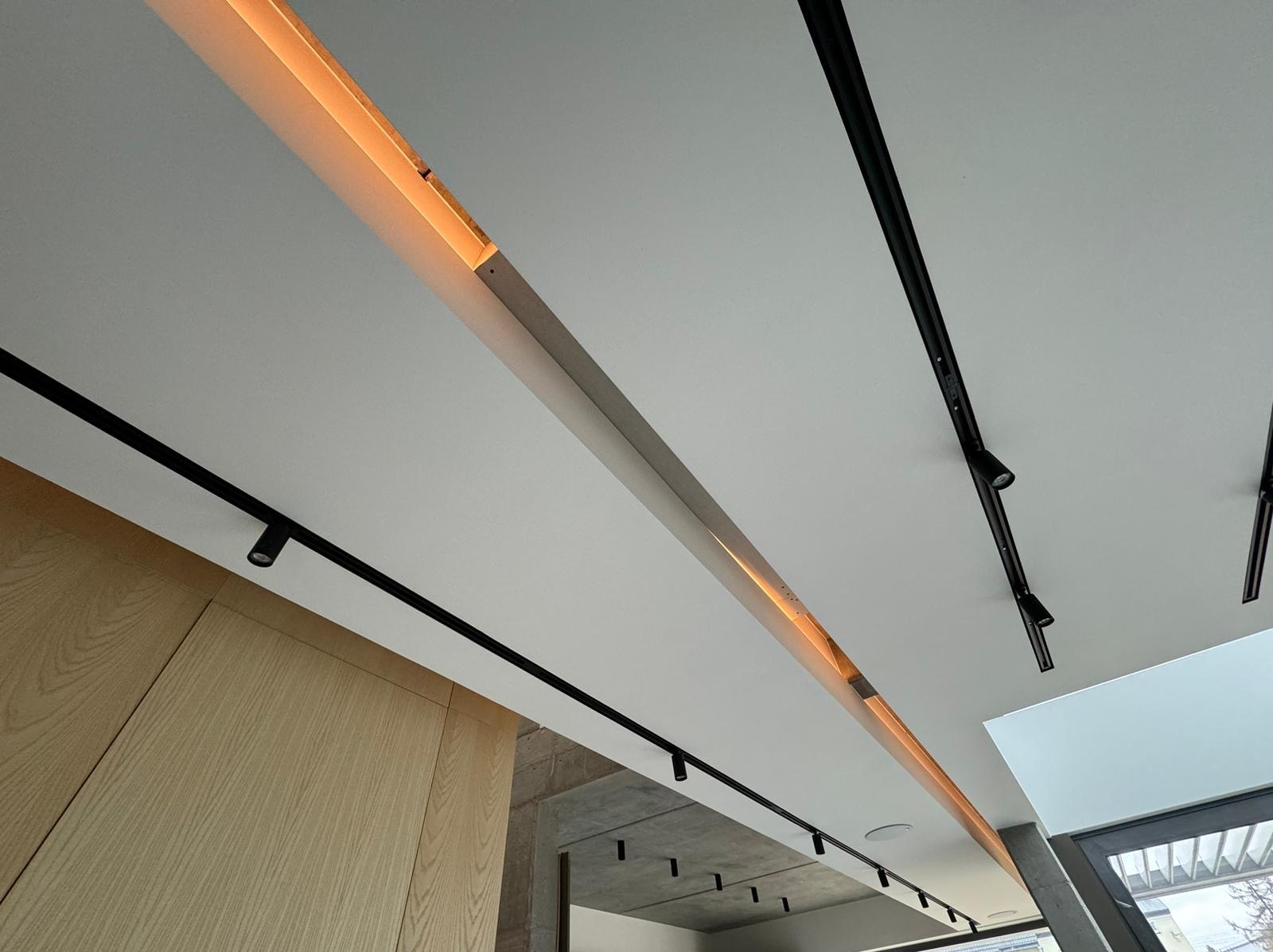 Nowoczesne oświetlenie sufitowe: czarne szyny z reflektorami punktowymi i podłużna oprawa z ciepłym światłem, widok z dołu.