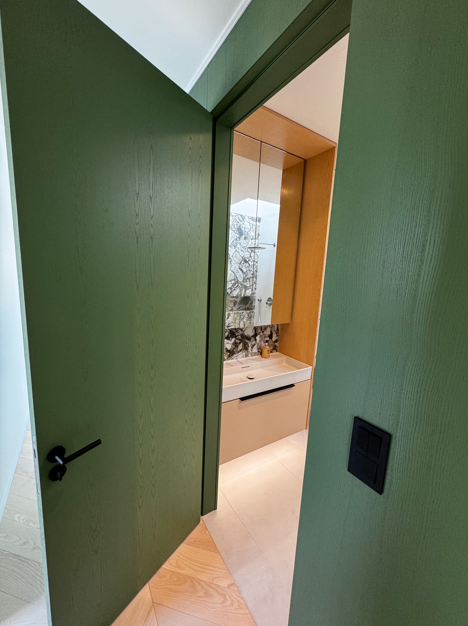 Otwór drzwiowy w odcieniach zieleni prowadzi do nowocześnie urządzonej łazienki z marmurową ścianą, beżową szafką pod umywalką i drewnianymi elementami wykończeniowymi.