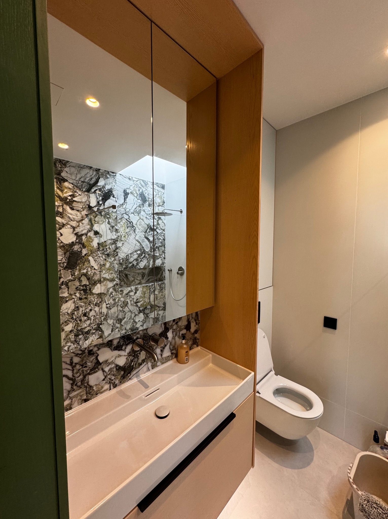 Nowoczesna łazienka z marmurową ścianą prysznicową odbijającą się w lustrze nad umywalką, wisząca toaleta z boku, minimalistyczny design.