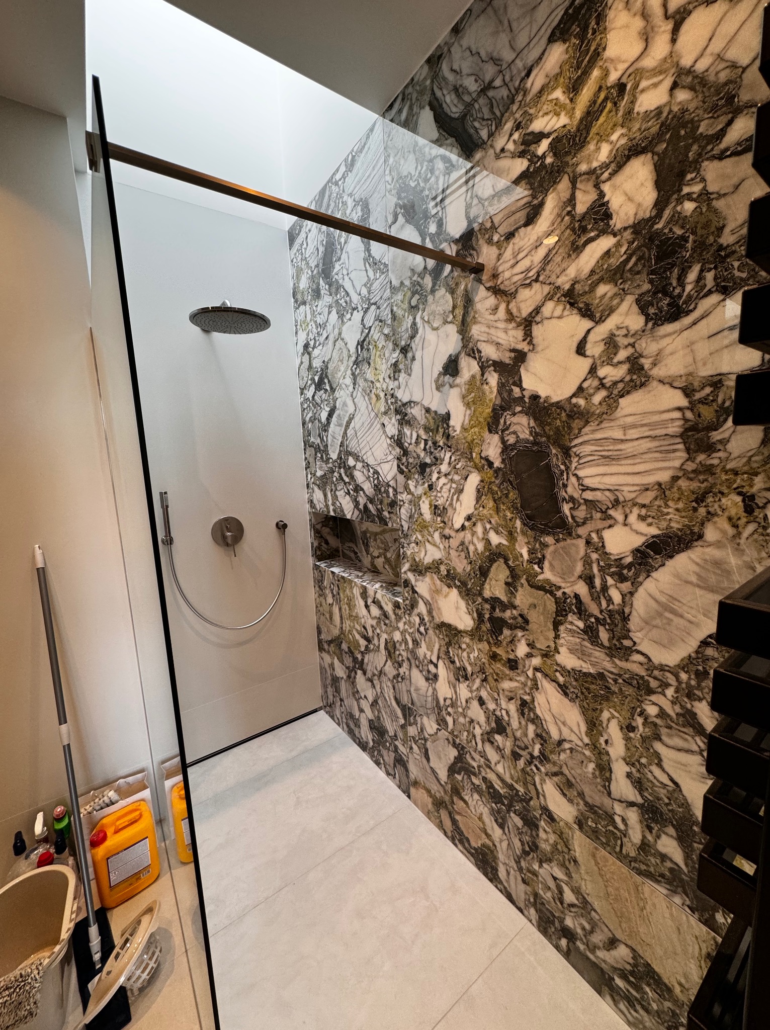 Nowoczesna kabina prysznicowa typu walk-in z marmurową ścianą w odcieniach bieli, szarości i brązu, szklana ścianka oddzielająca od reszty łazienki, widoczny zestaw prysznicowy i akcesoria...