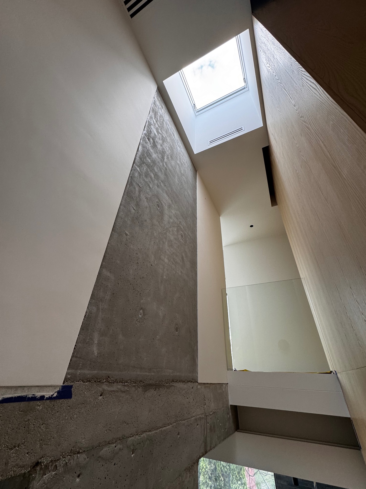 Wnętrze domu w trakcie wykańczania, surowe betonowe ściany kontrastują z gładkimi, pomalowanymi powierzchniami i drewnianą okładziną, widoczne okno dachowe wpuszczające naturalne światło.
