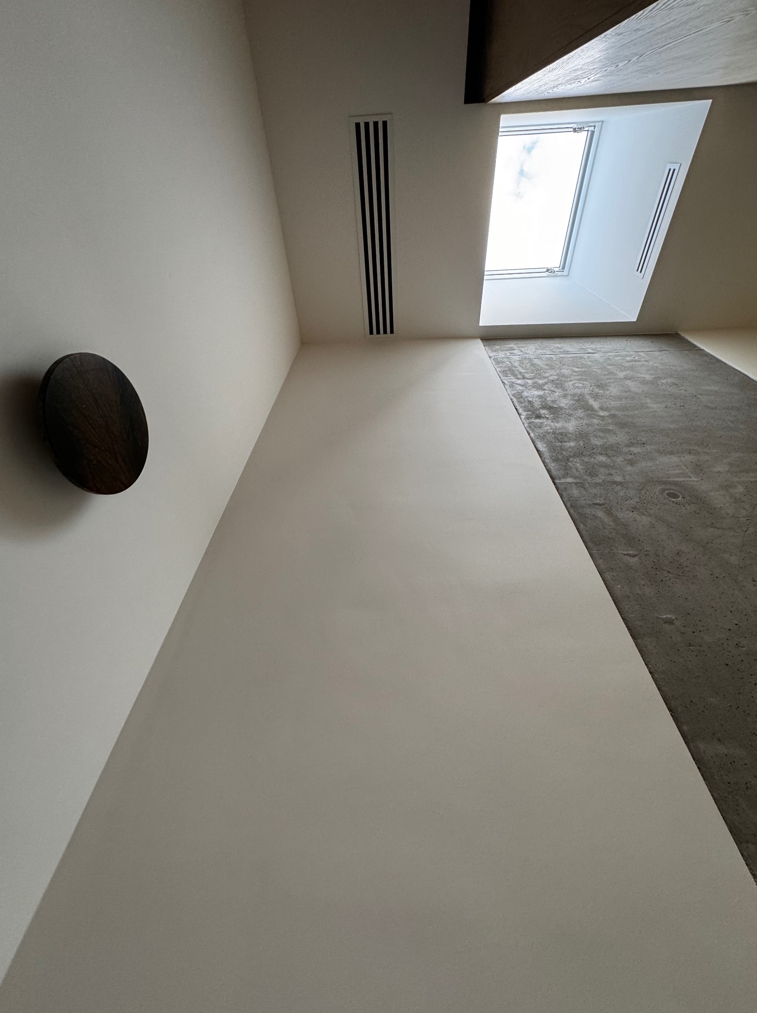 Minimalistyczne wnętrze w Poznaniu z widokiem na okno dachowe, szarą betonową podłogą i okrągłą drewnianą dekoracją ścienną.