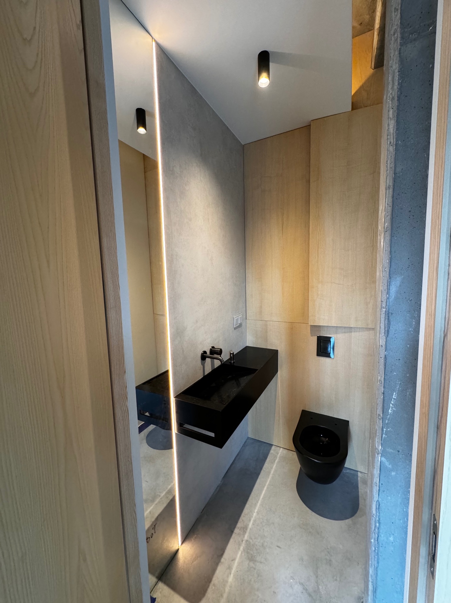 Nowoczesna łazienka z czarną armaturą, minimalistyczną umywalką i podwieszaną toaletą, oświetlenie LED wertykalne, surowy beton i drewniane panele ścienne.