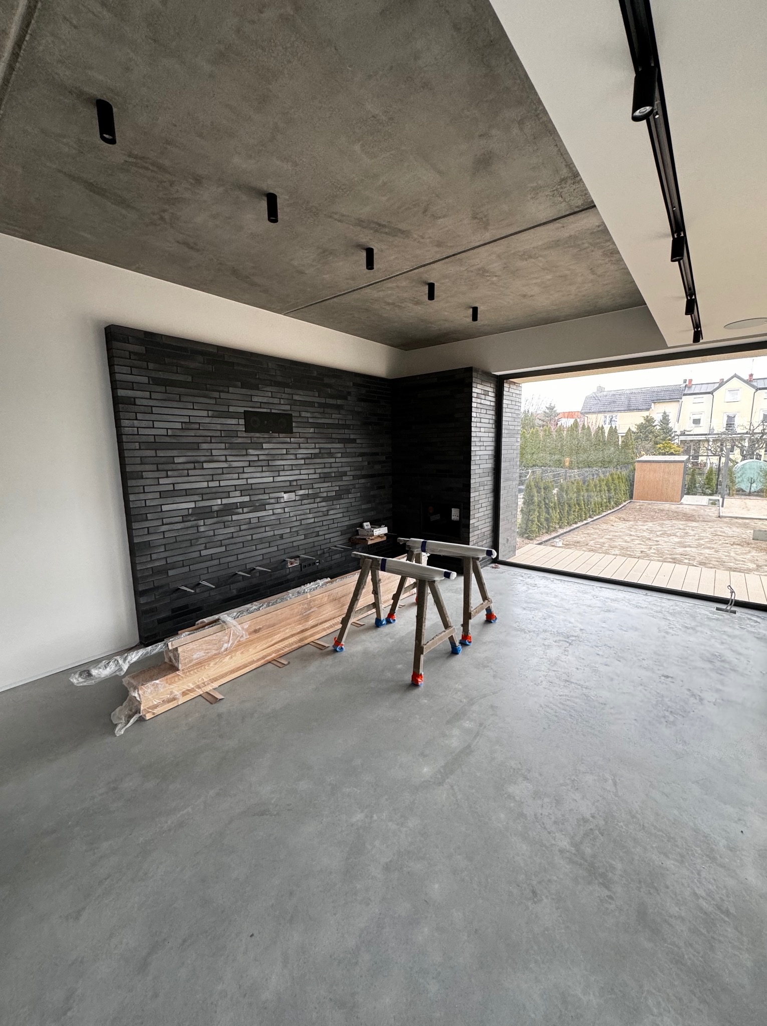 Wnętrze w trakcie wykańczania: ściana z czarnej cegły, betonowy sufit z reflektorami, duże okno z widokiem na ogród, drewniane deski w folii i kozły malarskie na szarym, betonowym podłożu.
