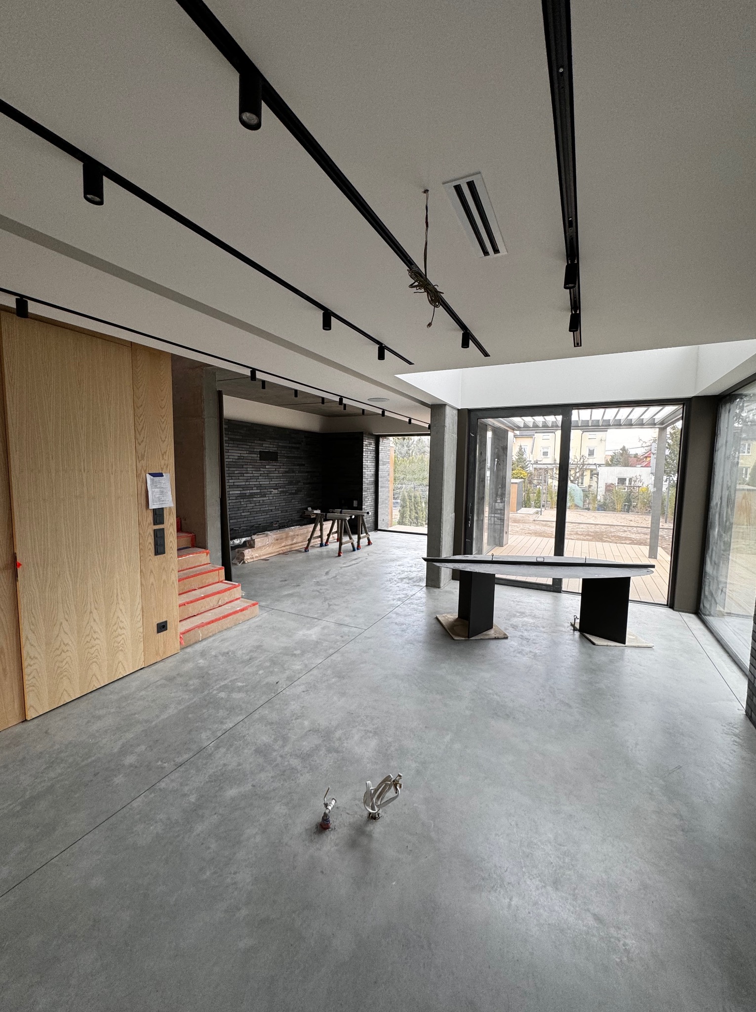 Wnętrze w trakcie wykańczania: surowy beton na podłodze, czarne szyny oświetleniowe na suficie, drewniana zabudowa, schody zabezpieczone folią, czarna ściana z cegły w głębi, widok na ogród przez...