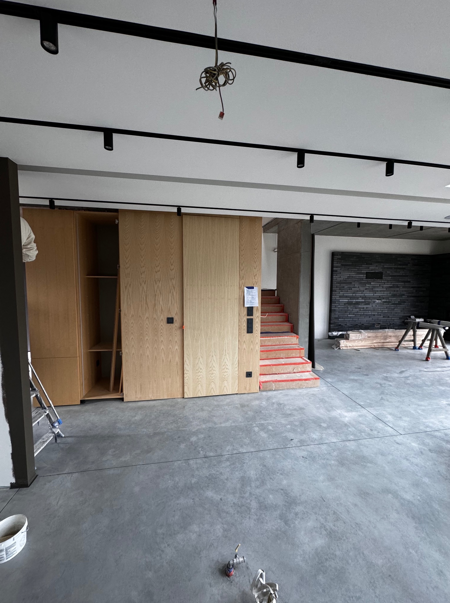 Wnętrze w trakcie wykończenia: drewniane szafy wnękowe z przesuwnymi drzwiami, zabezpieczone schody z betonowymi stopniami, surowa ściana z szarej cegły, widoczne narzędzia i kable oświetleniowe.