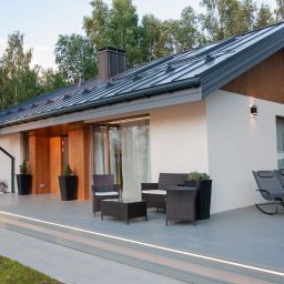 GES GROUP SPÓŁKA Z OGRANICZONĄ ODPOWIEDZIALNOŚCIĄ - Parterowy dom z białym tynkiem, ciemnoszarą blachą na dachu i drewnianymi elementami elewacji, z tarasem wyposażonym w meble ogrodowe i leżaki.