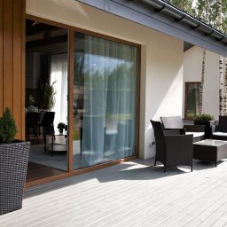 GES GROUP SPÓŁKA Z OGRANICZONĄ ODPOWIEDZIALNOŚCIĄ - Nowoczesny dom murowany z tarasem wyłożonym szarą deską kompozytową, widok na wnętrze przez przesuwne okno tarasowe, minimalistyczny zestaw mebli ogrodowych z rattanu.