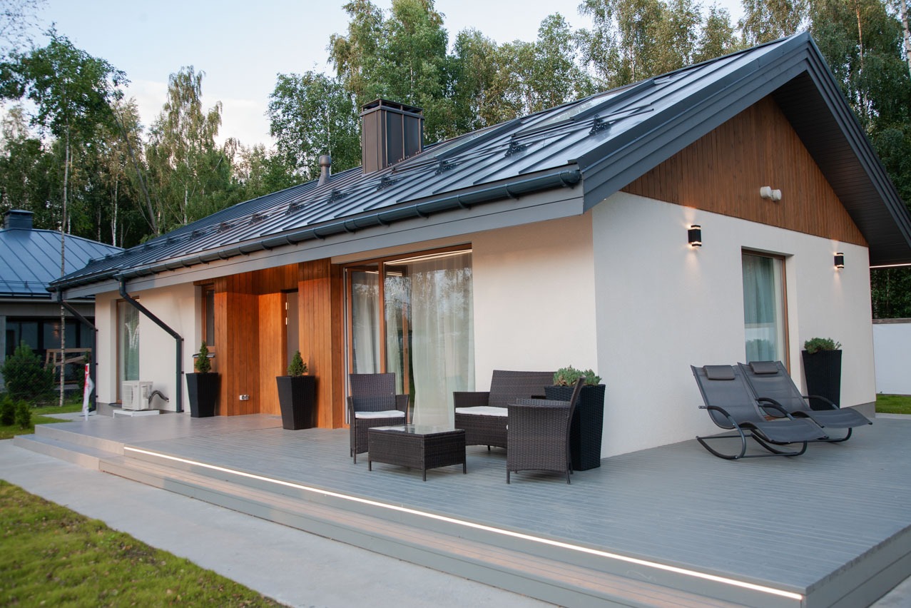 Parterowy dom z białym tynkiem, ciemnoszarą blachą na dachu i drewnianymi elementami elewacji, z tarasem wyposażonym w meble ogrodowe i leżaki.