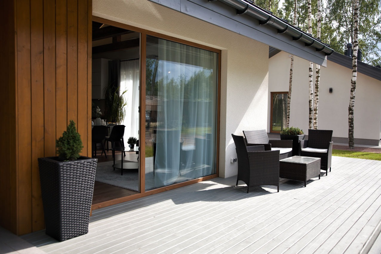 Nowoczesny dom murowany z tarasem wyłożonym szarą deską kompozytową, widok na wnętrze przez przesuwne okno tarasowe, minimalistyczny zestaw mebli ogrodowych z rattanu.