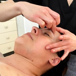 Ciałodusza - Kobieta leżąca na plecach podczas zabiegu na twarz, skupienie na masażu okolic czoła i brwi, widoczne dłonie masażysty w czarnym uniformie.