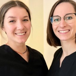 Ciałodusza - Portret dwóch uśmiechniętych kobiet w czarnych uniformach, prawdopodobnie pracownice salonu, jedna w okularach, obie z delikatnym makijażem, stojące na tle jasnej ściany.