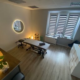 Ciałodusza - Jasne wnętrze gabinetu z łóżkiem do zabiegów, okrągłym lustrem z podświetleniem i drewnianą podłogą, widziane z góry.